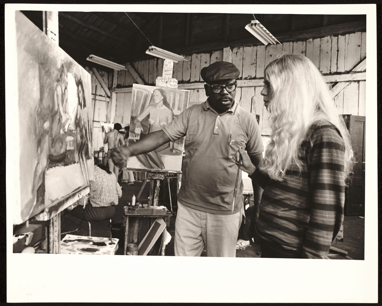 One person points to a painting on an easel while another person looks at the painting.