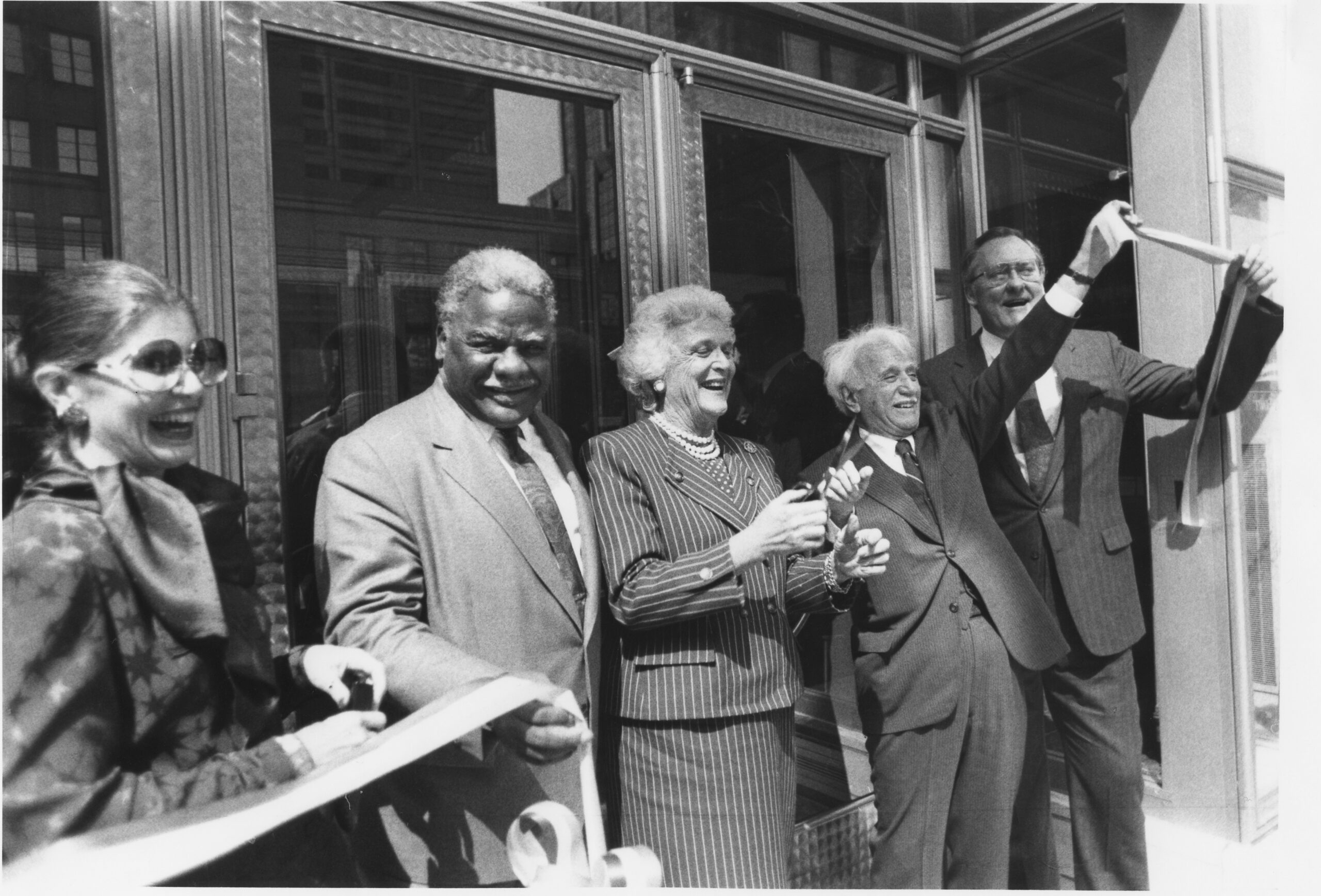 Five people stand in front of a set of doors. They all hold part of a ribbon that has been cut in two.