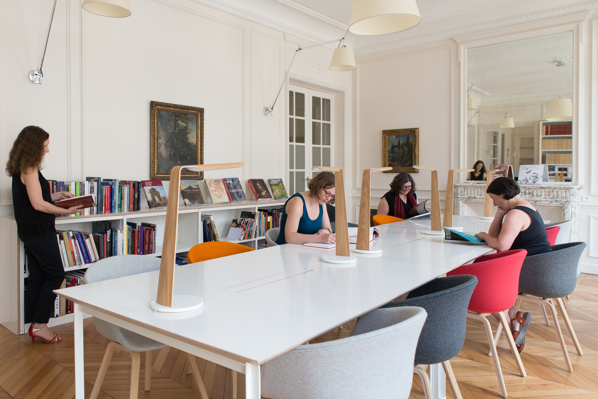 Three people reading and sitting at a long white table. A book shelf filled with books runs the length of the wall behind them.