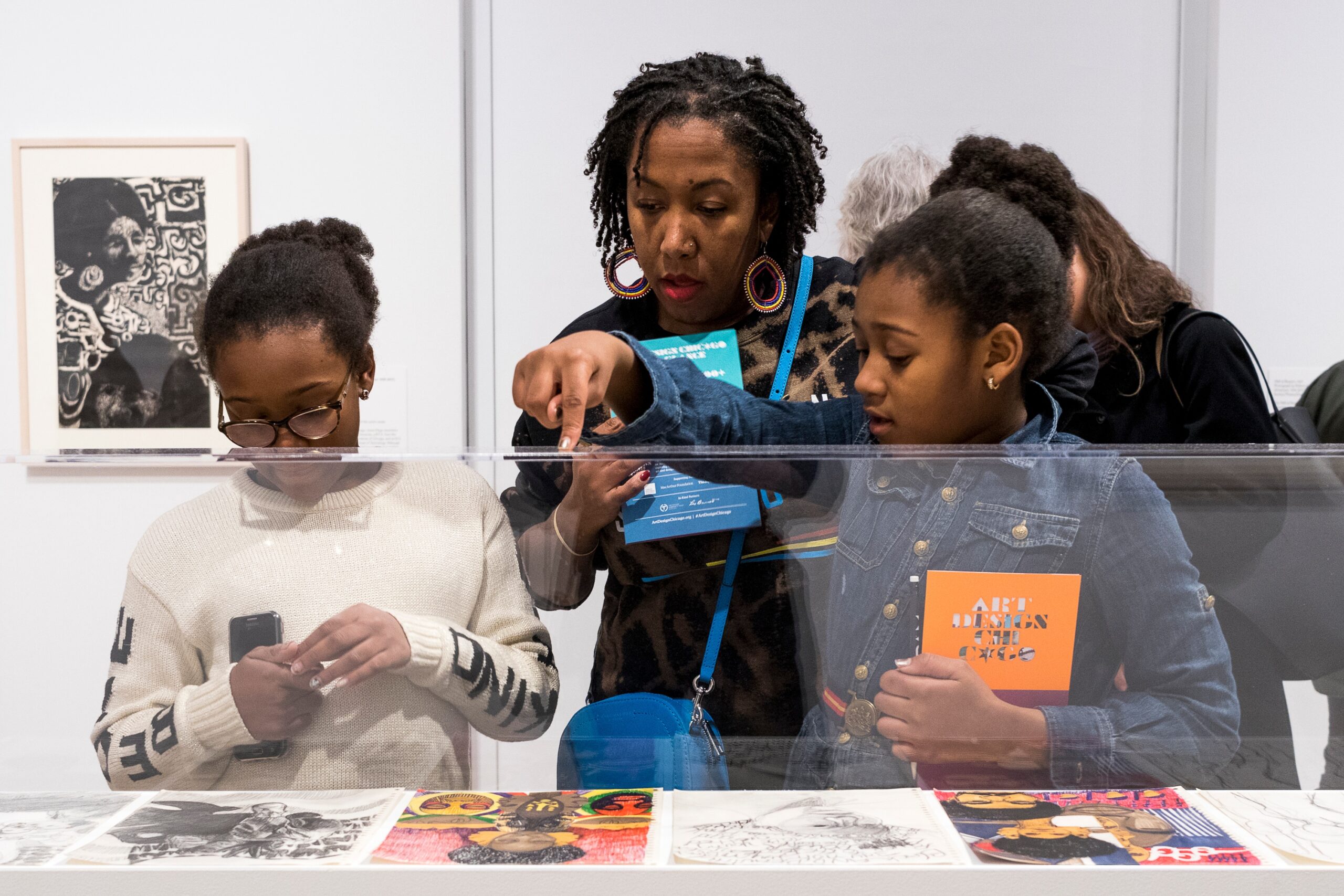 An adult and two children look into a case that contains flat, paper artworks. The adult and the child on the right hold booklets that say "Art Design Chicago."