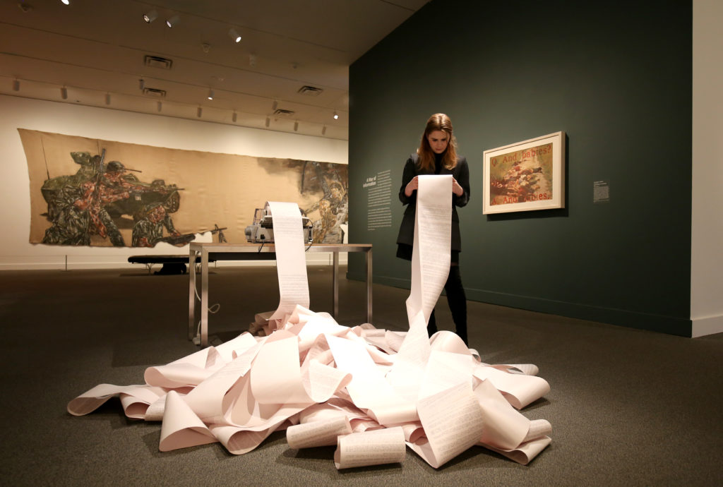 A woman stands holding a paper scroll. The scroll is unraveling and bunched up in a large pile on the floor of an exhibition space.