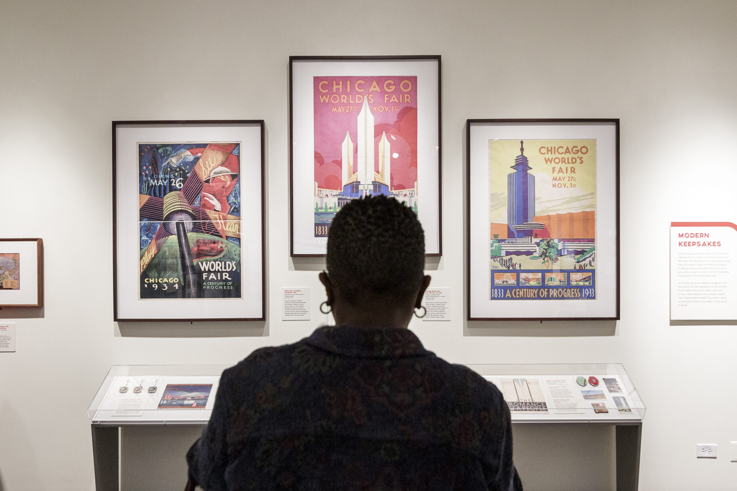 A person is standing, looking straight ahead, to a wall with three artworks hung side by side above a white table.