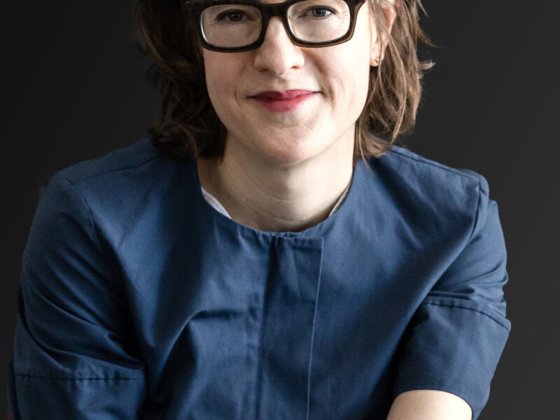 A woman with glasses and short brown hair is seated and smiling.