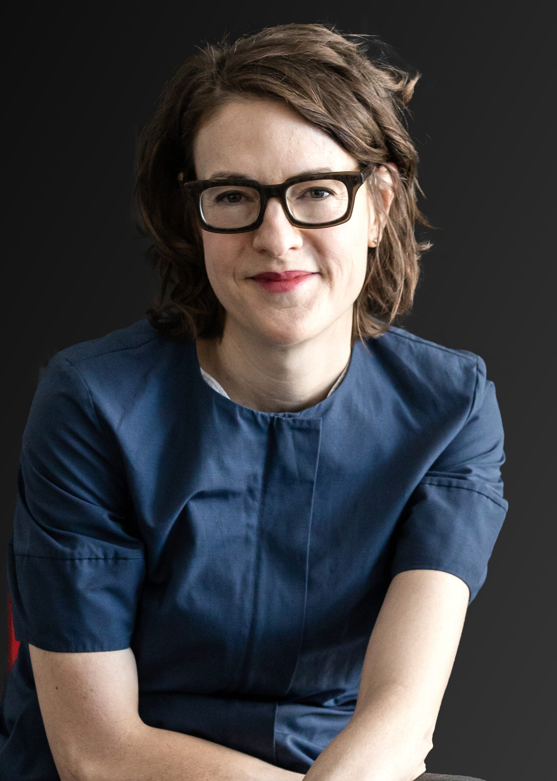 A woman with glasses and short brown hair is seated and smiling.