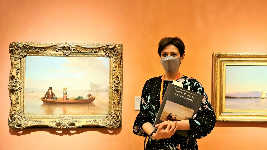 A woman holding a book and wearing a face mask stands in front of two paintings in gold frames. The gallery wall is painted a peach color.