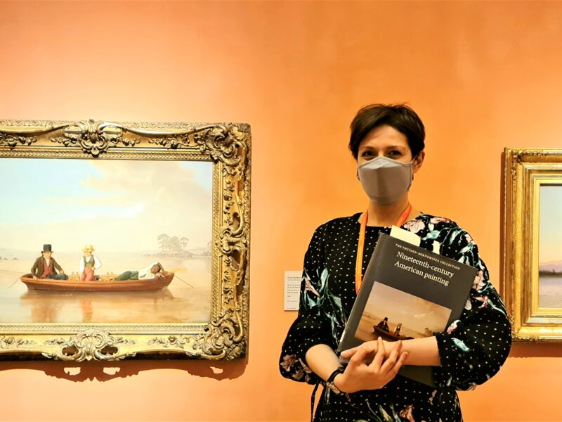 A woman holding a book and wearing a face mask stands in front of two paintings in gold frames. The gallery wall is painted a peach color.