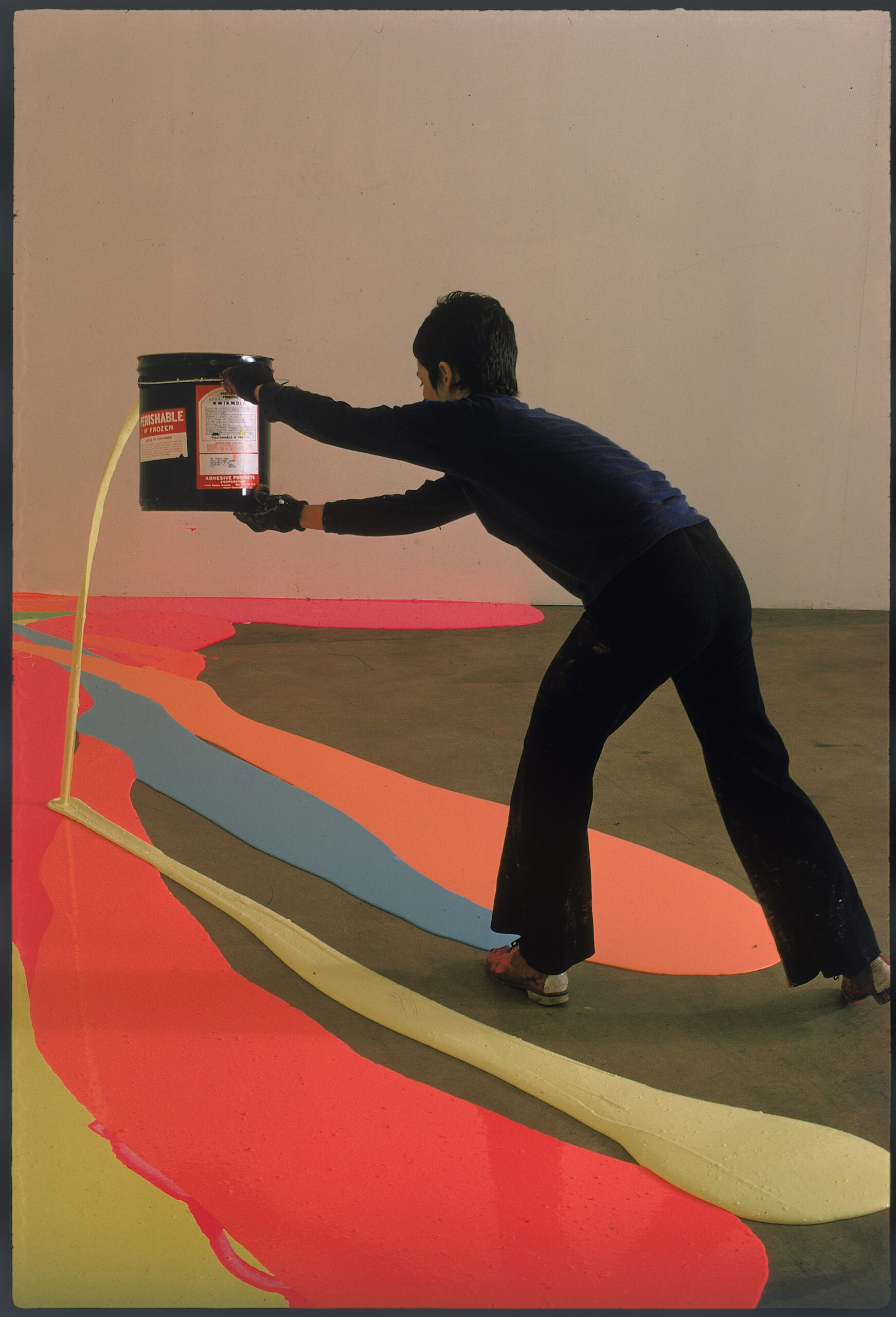 A woman dressed in black spilling paint from a paint can onto the floor, which already has paint spills in orange, blue, and yellow.
