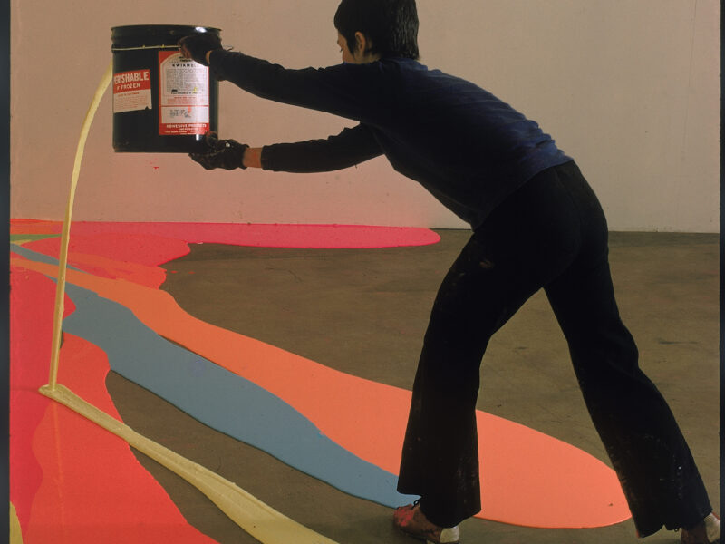 A woman dressed in black spilling paint from a paint can onto the floor, which already has paint spills in orange, blue, and yellow.
