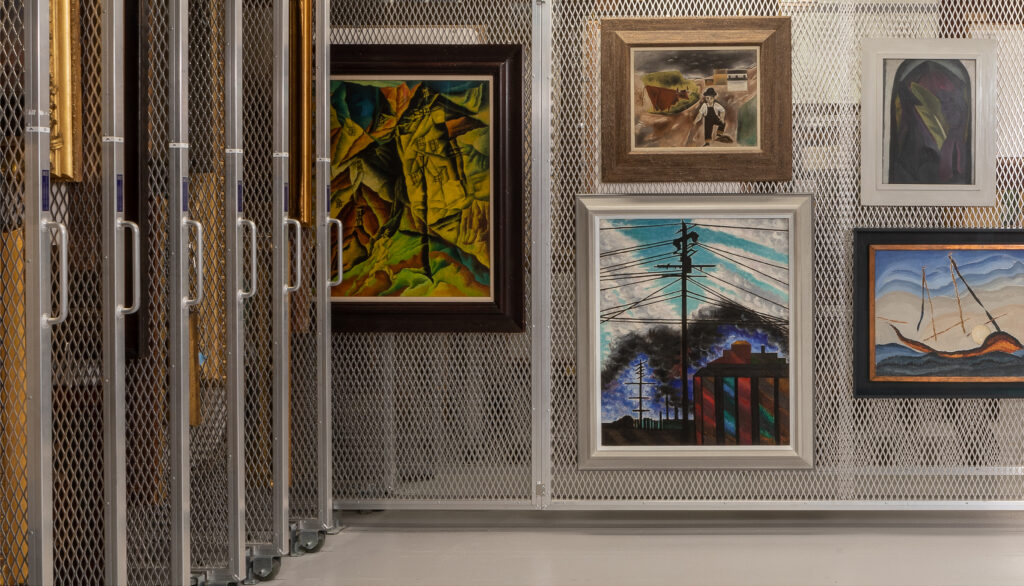Five framed paintings hang on a wire wall, pulled out from a row of moveable wire walls on wheels.
