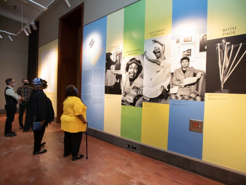 People viewing large photographs on a wall painted in blue, green, and yellow vertical stripes.