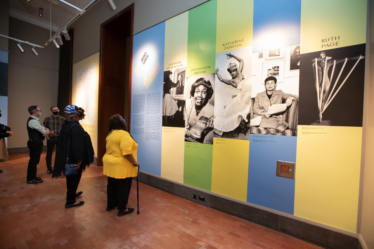 People viewing large photographs on a wall painted in blue, green, and yellow vertical stripes.