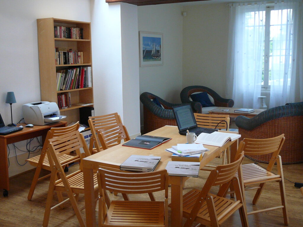 A room with a table and six empty chairs. The table holds books and papers.