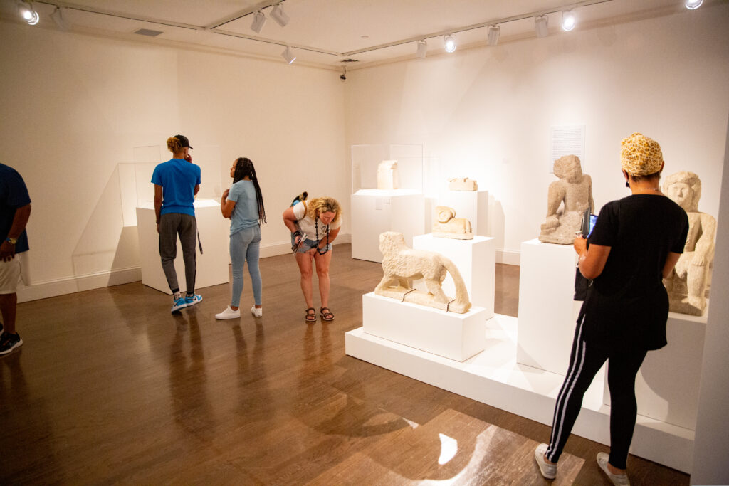 People viewing an exhibition of stone sculptures. The sculptures are white stones in various shapes resembling animals and seated human figures.