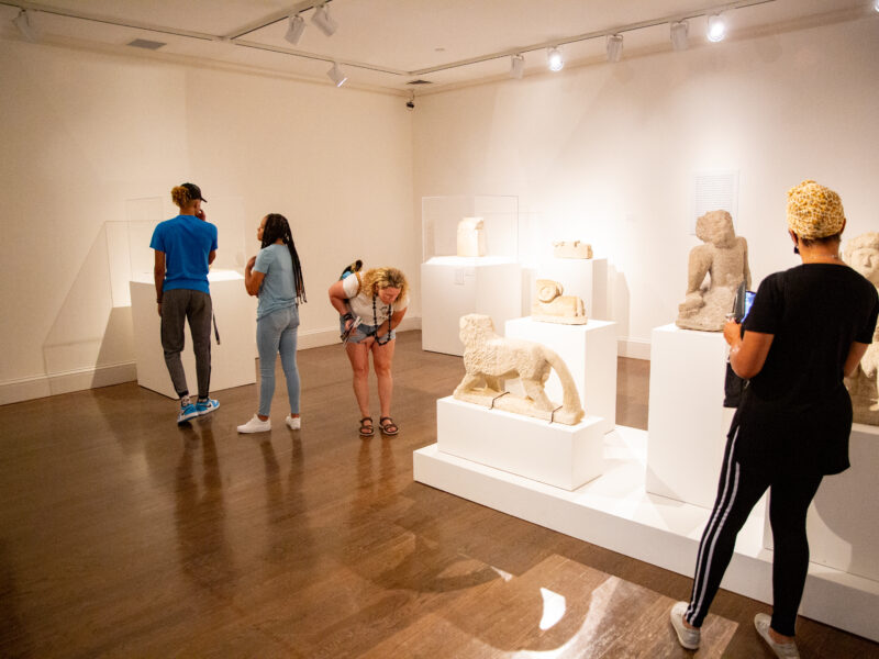 People viewing an exhibition of stone sculptures. The sculptures are white stones in various shapes resembling animals and seated human figures.