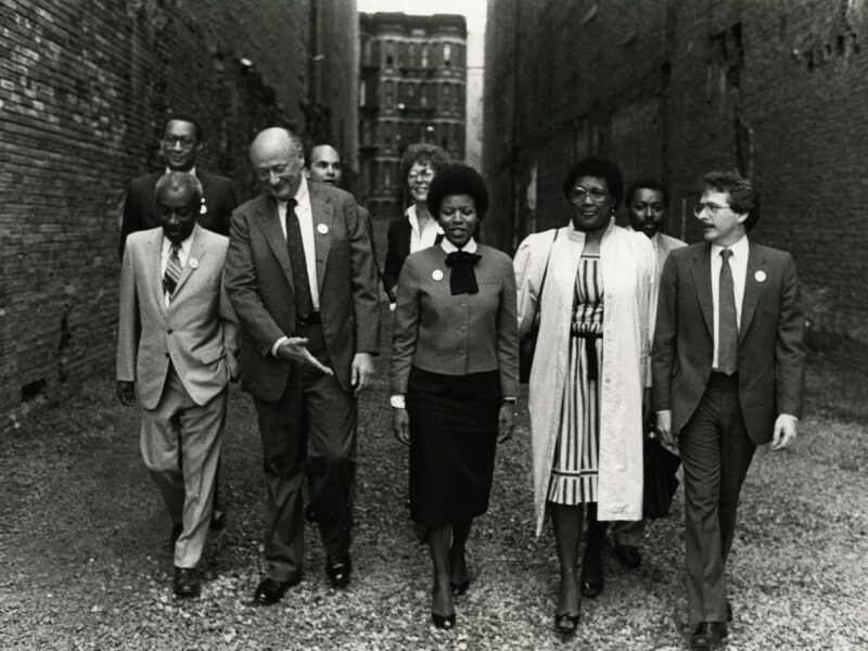 A black and white photo of a group of people dressed in business attire walking together between two buildings in New York City.