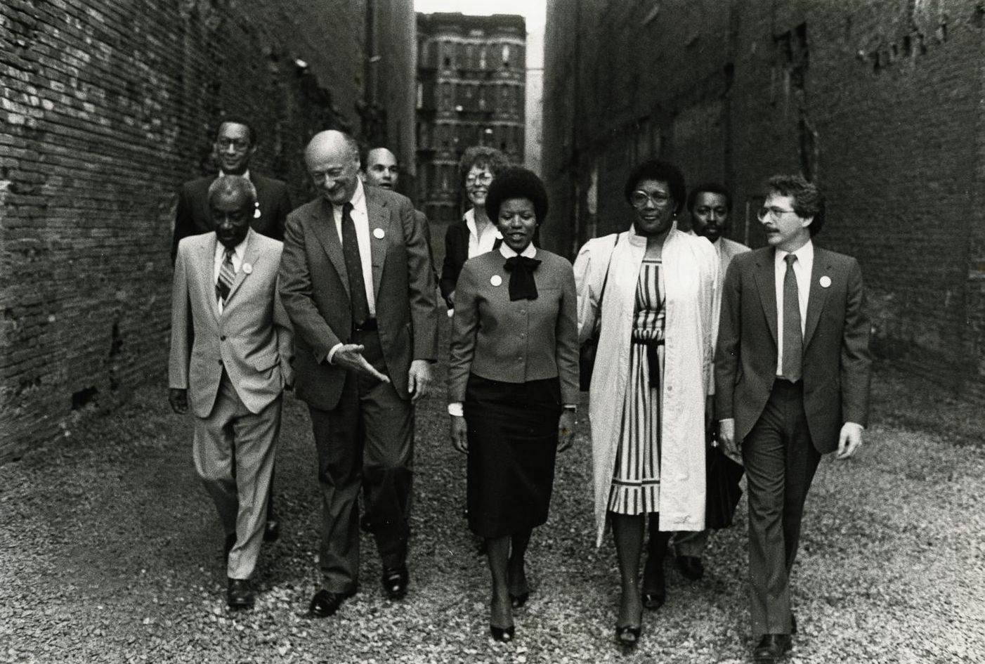 A black-and-white photograph of a group of people dressed in business attire walking together between two buildings in New York City.