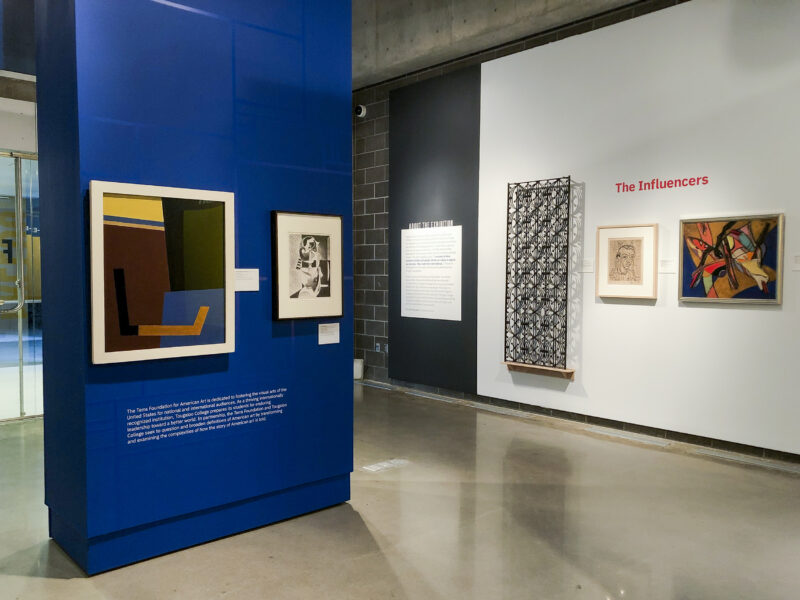 Installation photograph of an exhibition. Two paintings hang on a blue wall on the left side of the image, and three artworks hang on a white wall on the right side of the image.