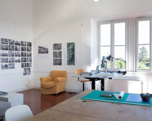 Artist studio space with grid of black-and-white photographs on one wall, a table in the center, and a window open to green space outside.
