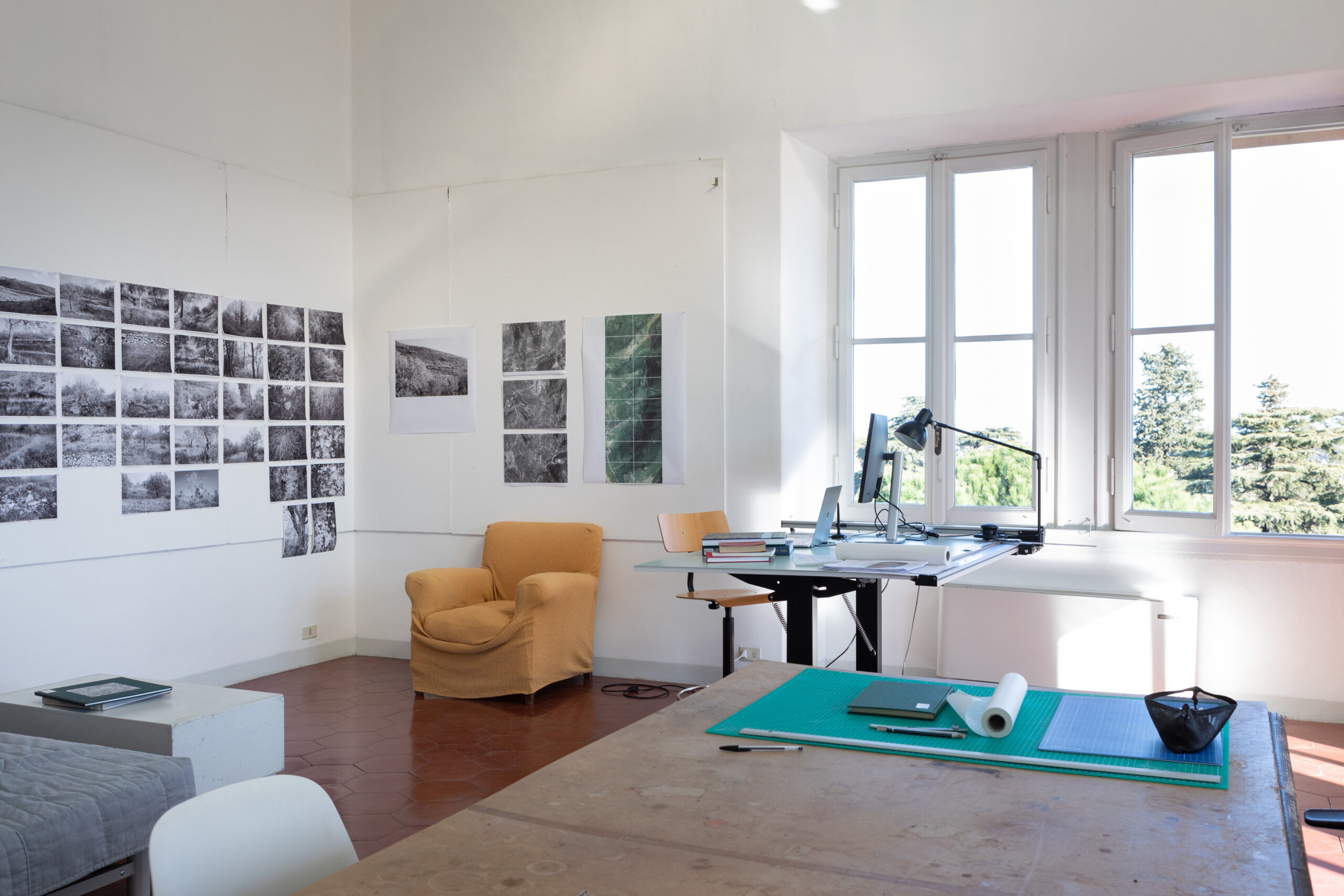 Artist studio space with grid of black-and-white photographs on one wall, a table in the center, and a window open to green space outside.