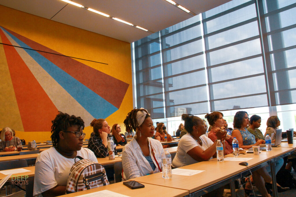 A diverse group of people seated at long tables in a classroom setting. They are paying attention to someone or something outside the frame.