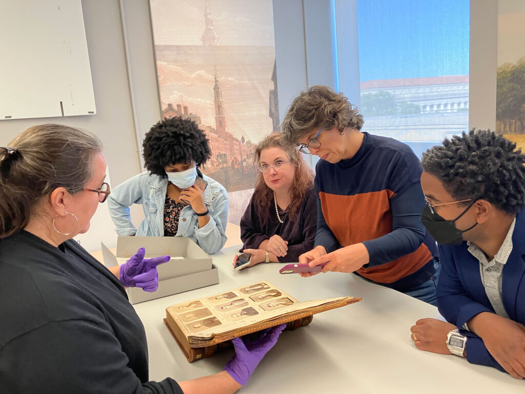 Five people gather around an open book featuring nine antique photographs of individuals. One person holds the book open with gloved hands and speaks to the group while another person takes a photograph with their cellphone of the open page.