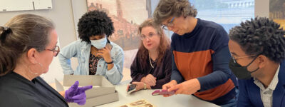 Five people gather around an open book featuring nine antique photographs of individuals. One person holds the book open with gloved hands and speaks to the group while another person takes a photograph with their cellphone of the open page.