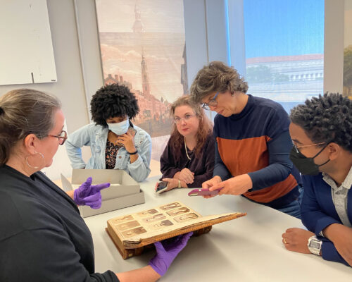 Five people gather around an open book featuring nine antique photographs of individuals. One person holds the book open with gloved hands and speaks to the group while another person takes a photograph with their cellphone of the open page.