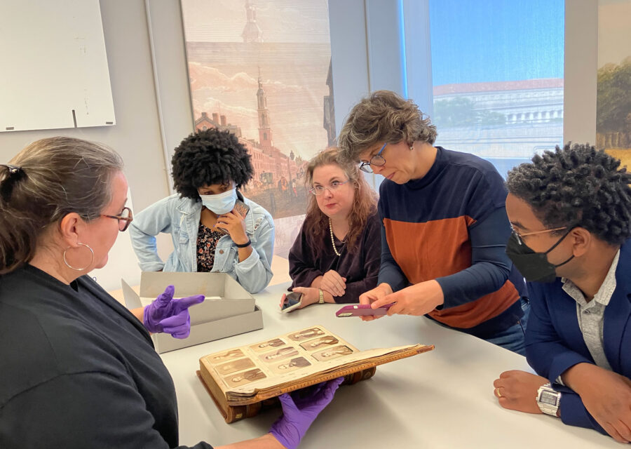 Five people gather around an open book featuring nine antique photographs of individuals. One person holds the book open with gloved hands and speaks to the group while another person takes a photograph with their cellphone of the open page.