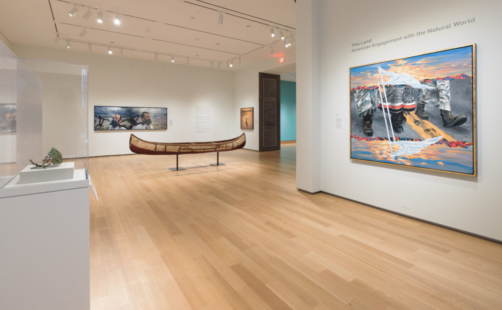 A photograph of a brightly lit museum gallery featuring three large contemporary paintings, a canoe, and an object in a case.