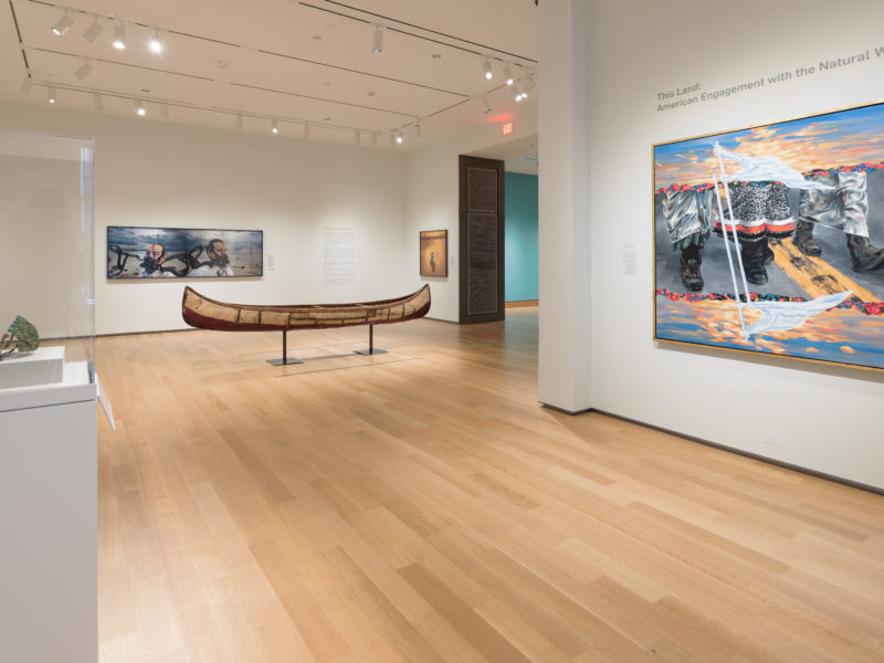 A photograph of a brightly lit museum gallery featuring three large contemporary paintings, a canoe, and an object in a case.