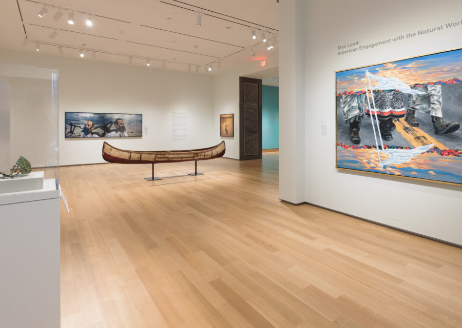 A photograph of a brightly lit museum gallery featuring three large contemporary paintings, a canoe, and an object in a case.