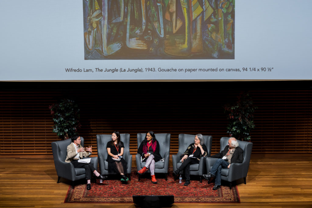 Five speakers are seated on a brightly lit stage against a digital screen featuring an abstract painting by Wifredo Lam. One of the speakers is talking and gesturing with his hands. The other four speakers watch while they listen to the person speak.