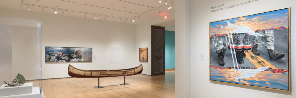 A photograph of a brightly lit museum gallery featuring three large contemporary paintings, a canoe, and an object in a case.