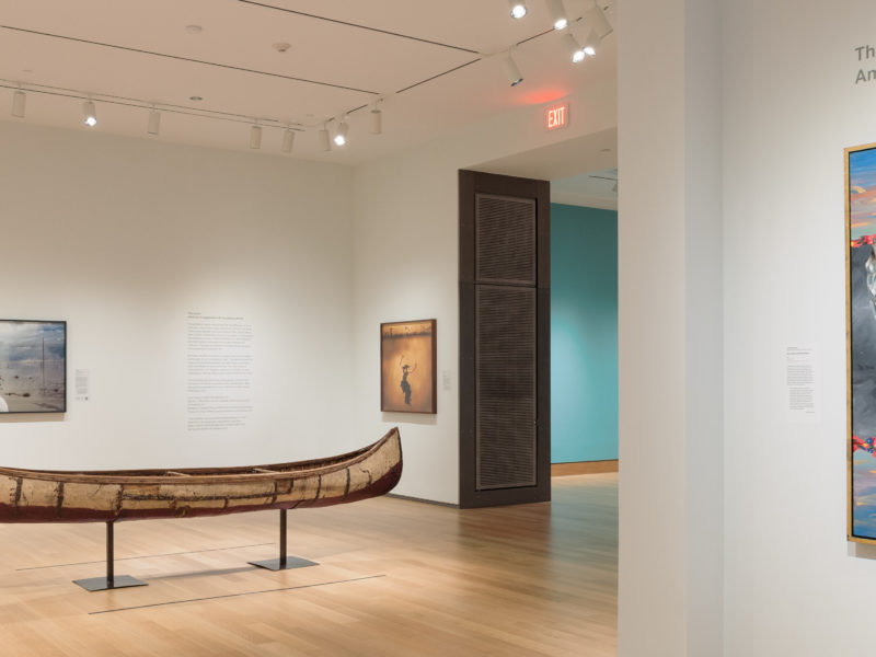 A photograph of a brightly lit museum gallery featuring three large contemporary paintings, a canoe, and an object in a case.