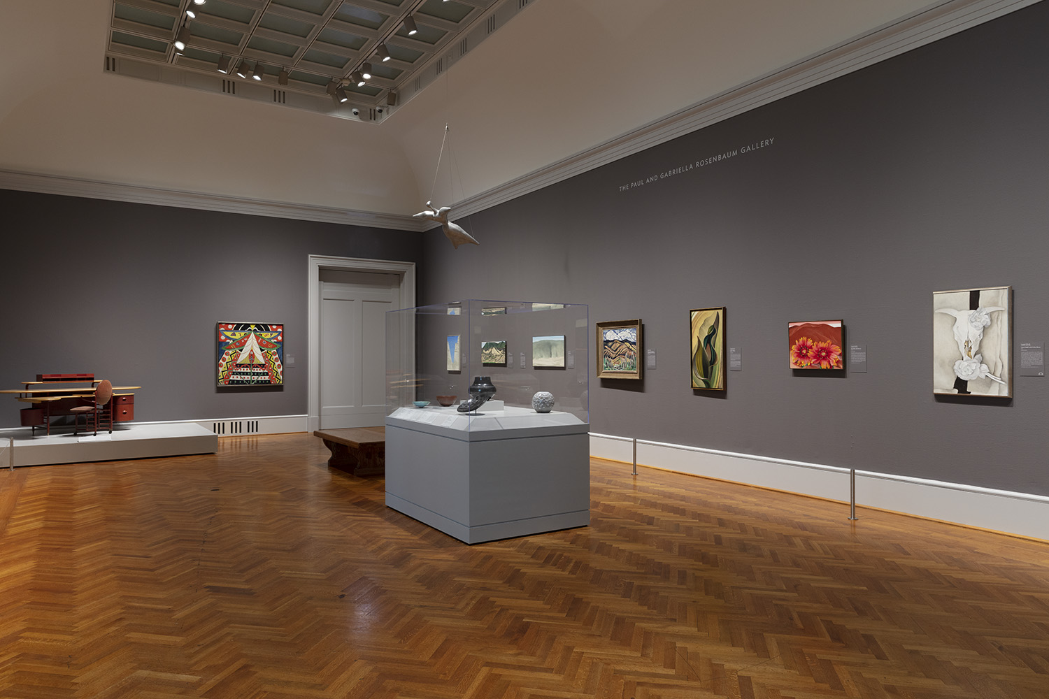 A photograph of a museum gallery featuring eight paintings of various sizes, a display case of ceramic vessels, and a desk.