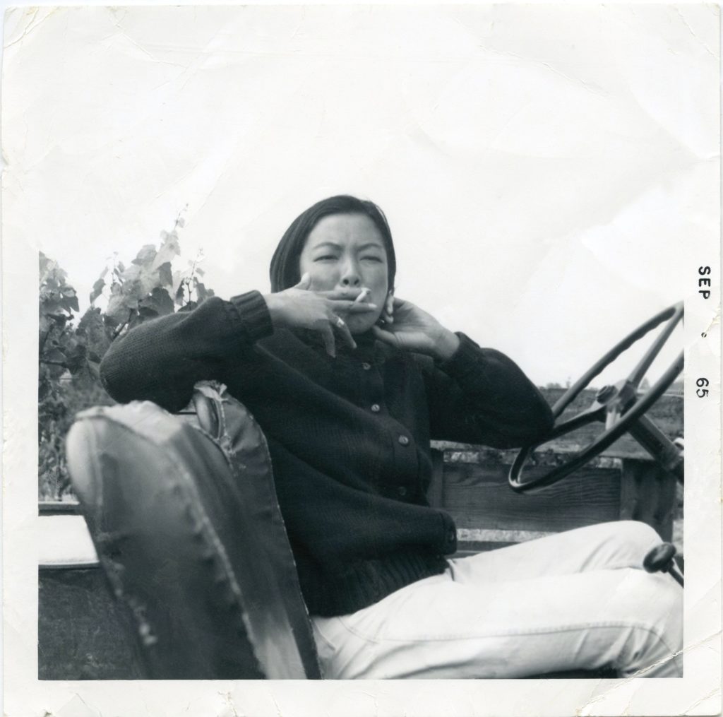 Woman seated in the driver’s seat of a car with no roof. She is smoking a cigarette and looking at the viewer.