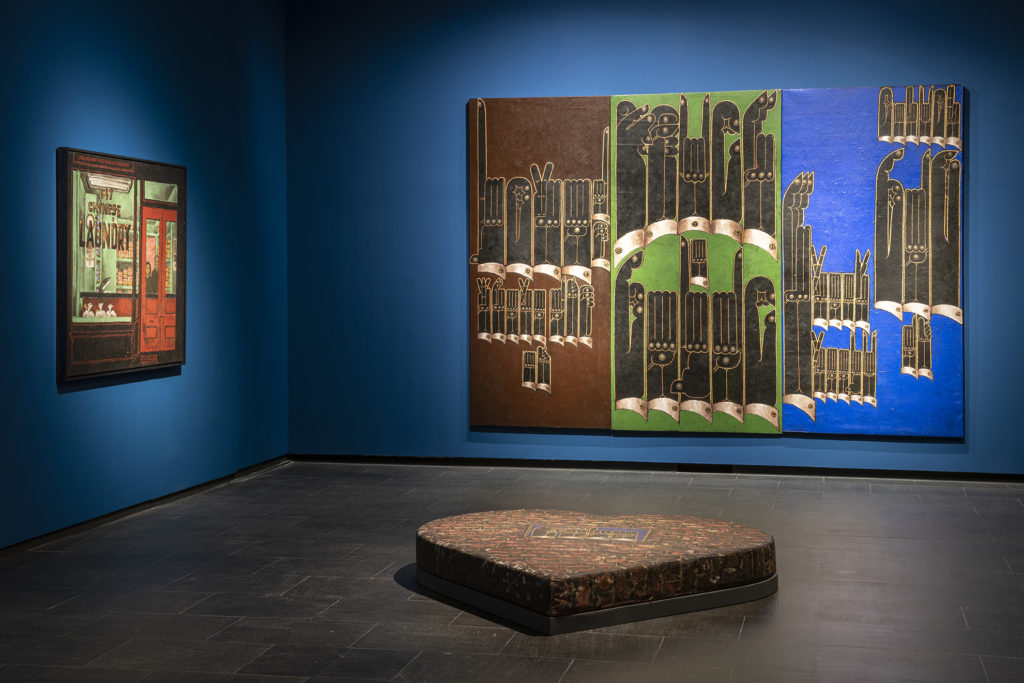 Two paintings hanging in a gallery with royal blue walls. A sculpture about 12 inches tall and a few feet wide in the shape of a heart is in the center of the gallery.