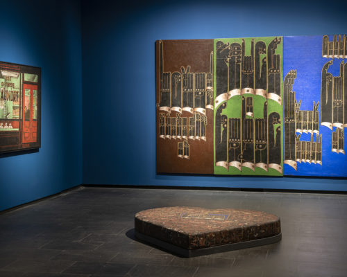 Two paintings hanging in a gallery with royal blue walls. A sculpture about 12 inches tall and a few feet wide in the shape of a heart is in the center of the gallery.