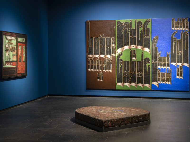 Two paintings hanging in a gallery with royal blue walls. A sculpture about 12 inches tall and a few feet wide in the shape of a heart is in the center of the gallery.