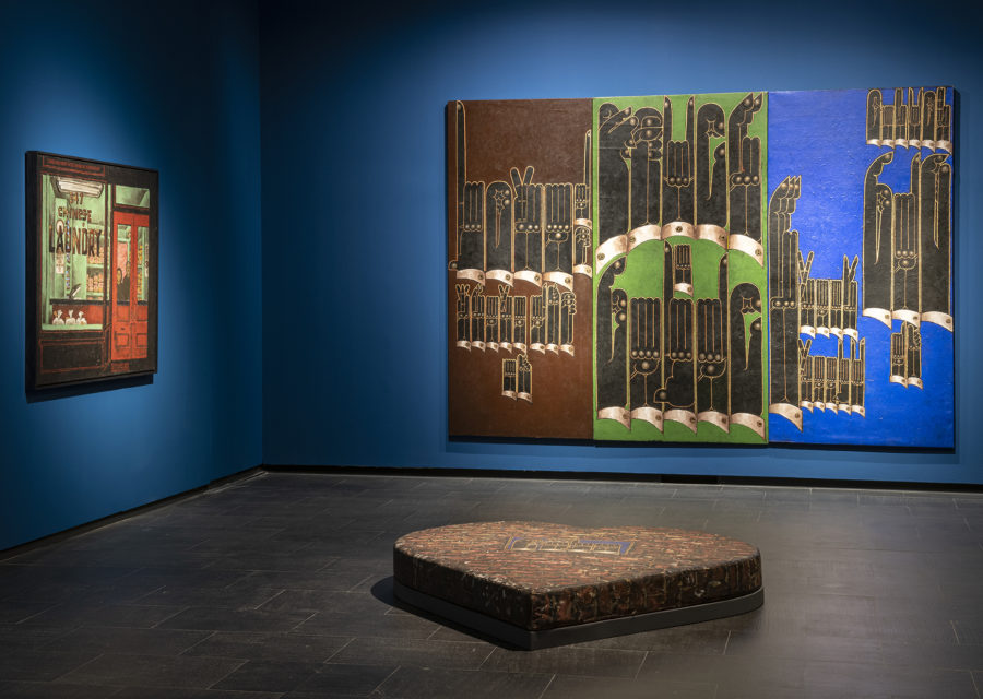 Two paintings hanging in a gallery with royal blue walls. A sculpture about 12 inches tall and a few feet wide in the shape of a heart is in the center of the gallery.