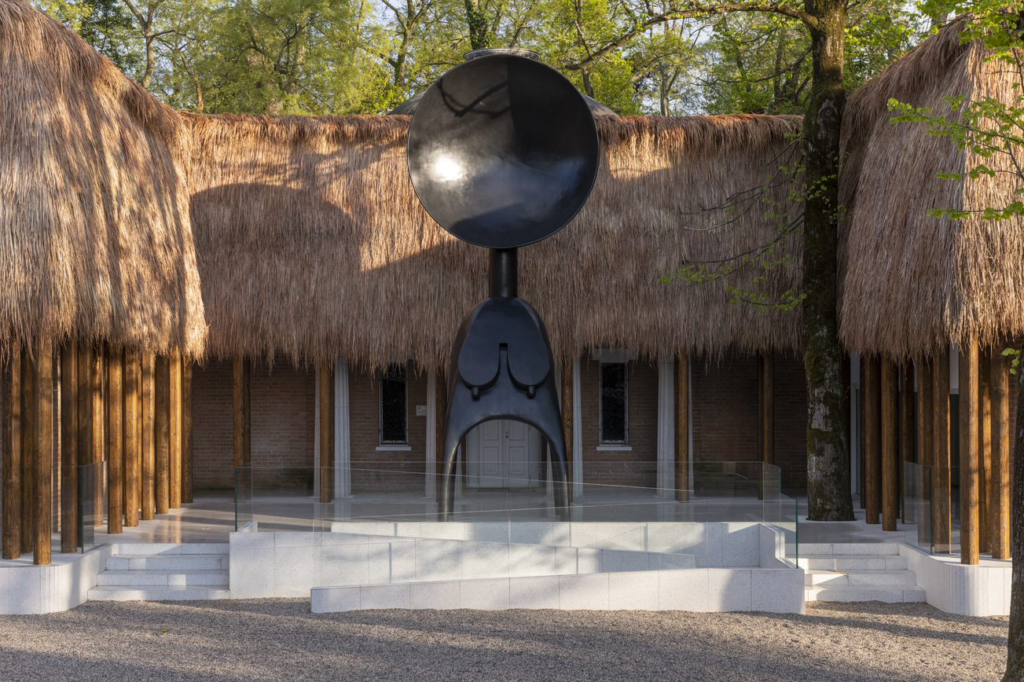 Abstract sculpture of a female form located in front of a building with a thatched roof.