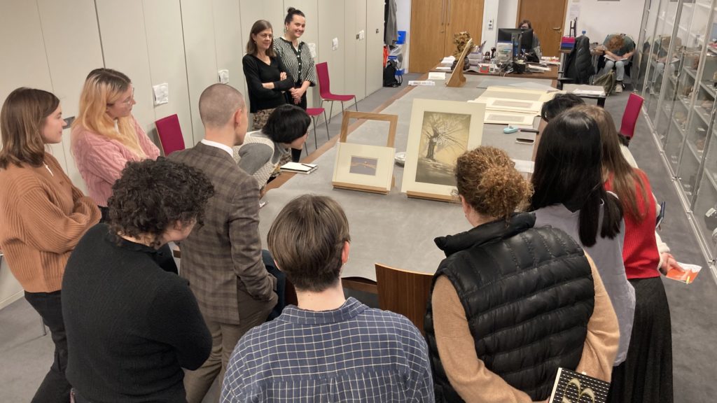 A group of people looking at two prints set upon small easels atop a long table.