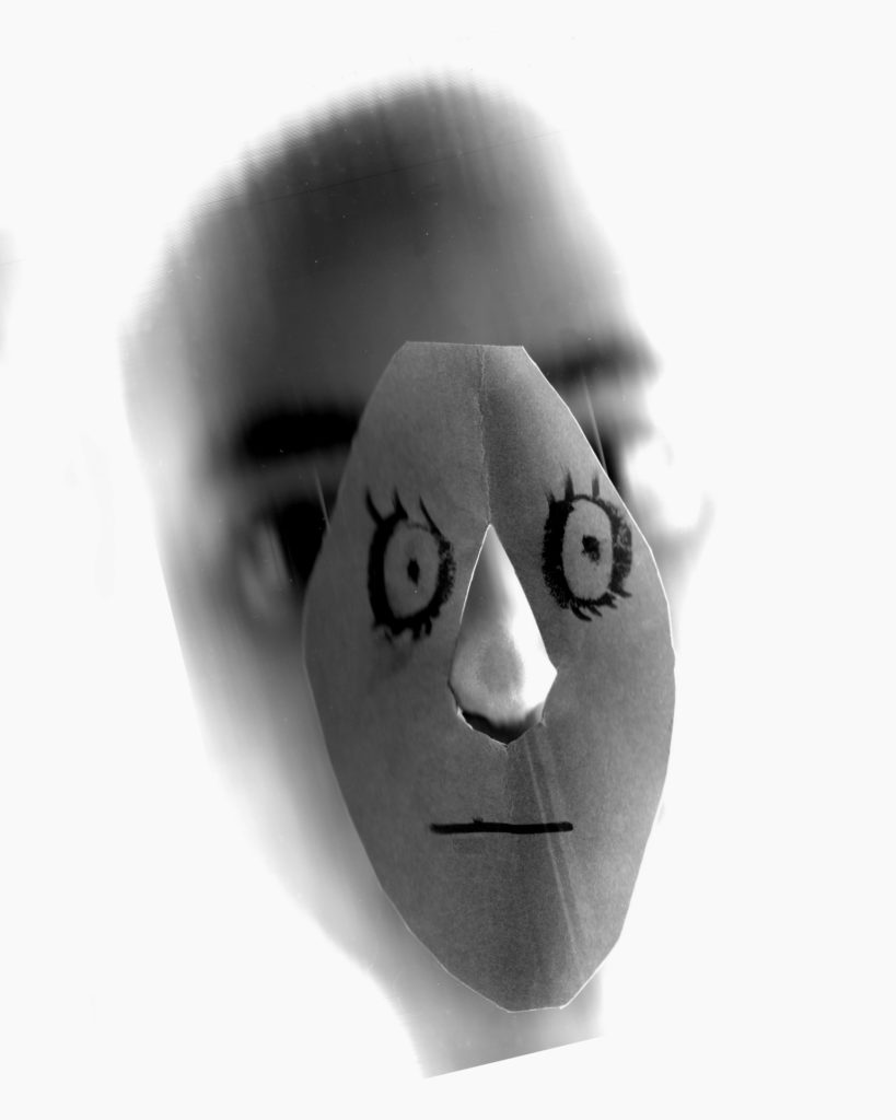 A black-and-white image of an obscured face with a paper cutout set upon a nose. The cutout has drawn-on eyes and a mouth.