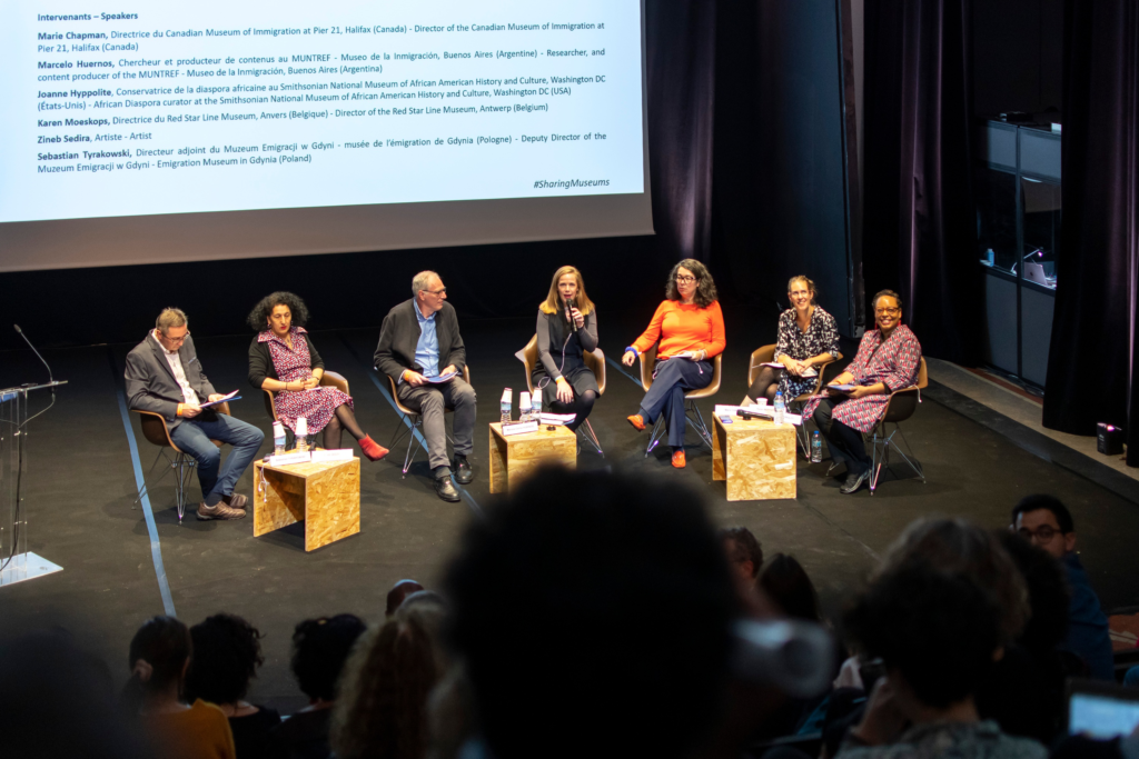 A photograph looking down toward an auditorium stage on which seven people are seated. One person on the stage holds a microphone and addresses the room.