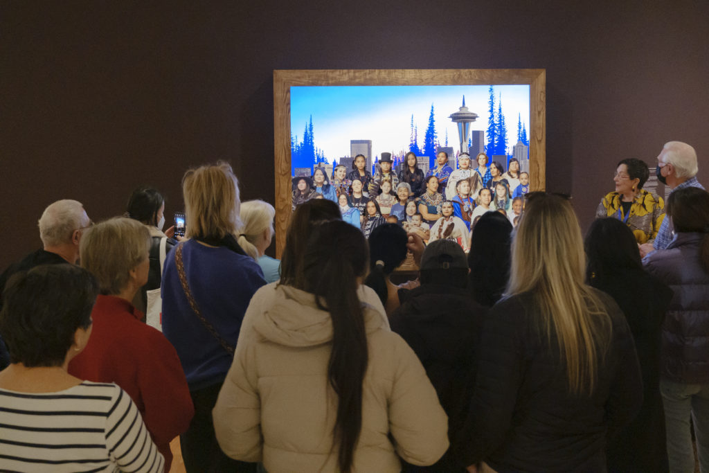 A group of people look at a painting while a person addresses them, talking about the painting. The painting features a group of people set against a city skyline.