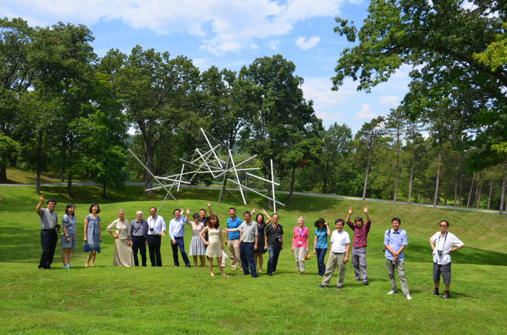 A group of people standing on a green lawn with a metal sculpture and trees behind them.