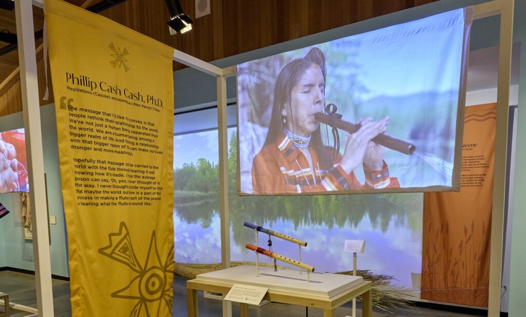 Exhibition installation image featuring a Phillip Cash, Ph.D., (Cayuse, Nez Perce) playing a woodwind instrument.