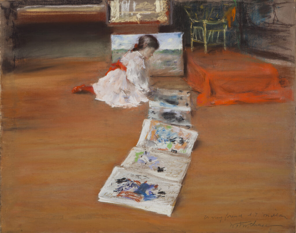 A girl in a white dress sits on a wood floor looking at a a book of Japanese prints unfolded.