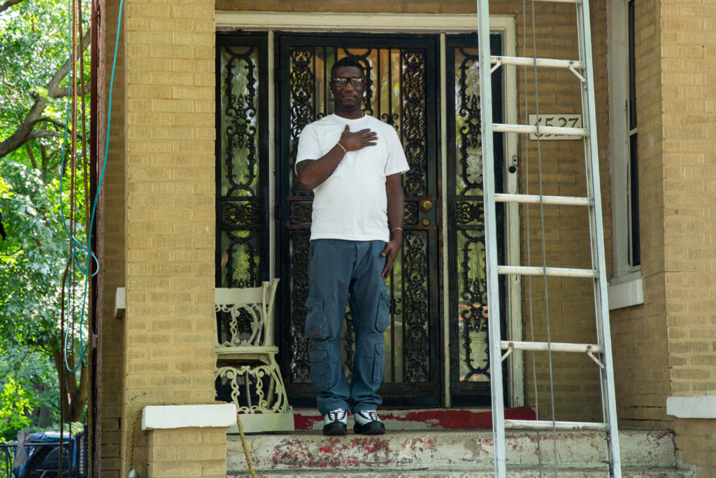 A person standing on the front porch of a house, with his hand on his chest, next to a ladder.