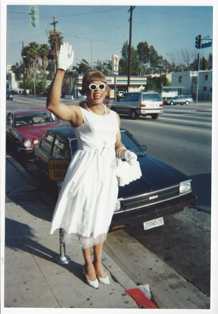 A picture of a person in a white dress waving, standing in front of a black car, on a sidewalk.